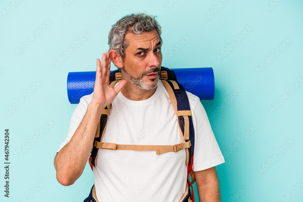 Middle age hiker caucasian man isolated on blue background  trying to listening a gossip.