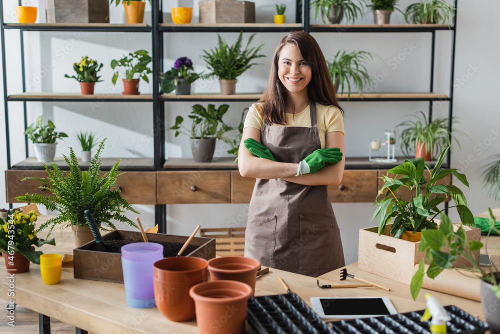 Smiling florist in gloves looking at camera near digital tablet and gardening tools in flower shop Smiling florist in gloves looking at camera near digital tablet and gardening tools in flower shop