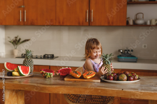 Healthy food. happy child girl and a lot of fruit. Cute little girl holding pineapple while sitting nar wooden table in home kitchen photo