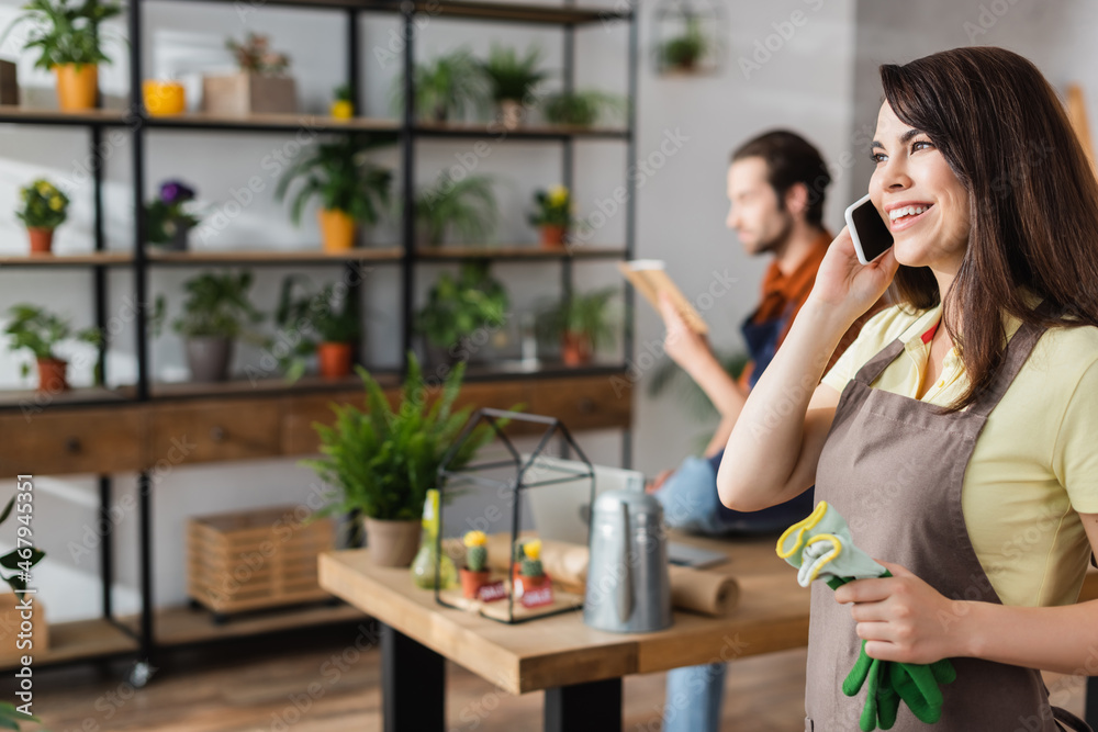 Smiling seller holding gardening gloves and talking on smartphone in flower shop Smiling seller holding gardening gloves and talking on smartphone in flower shop