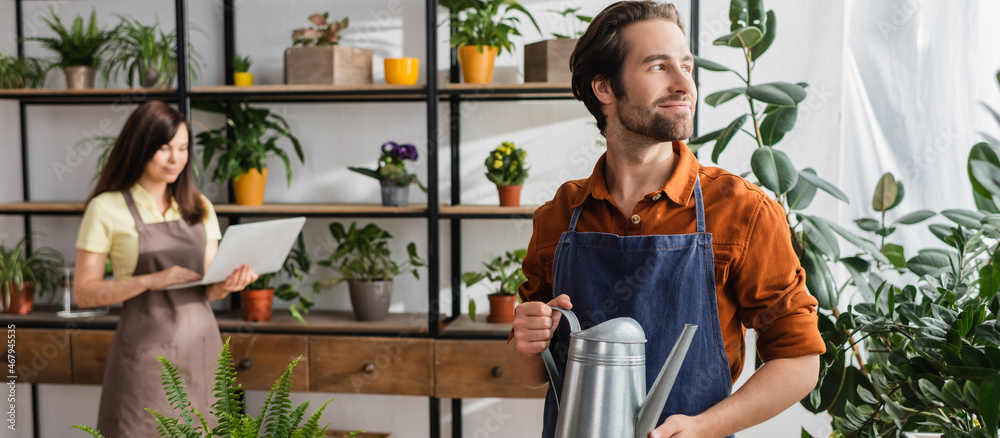 Seller holding watering can near plants and blurred colleague in shop, banner