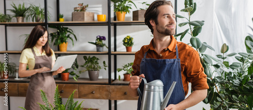 Seller holding watering can near plants and blurred colleague in shop, banner