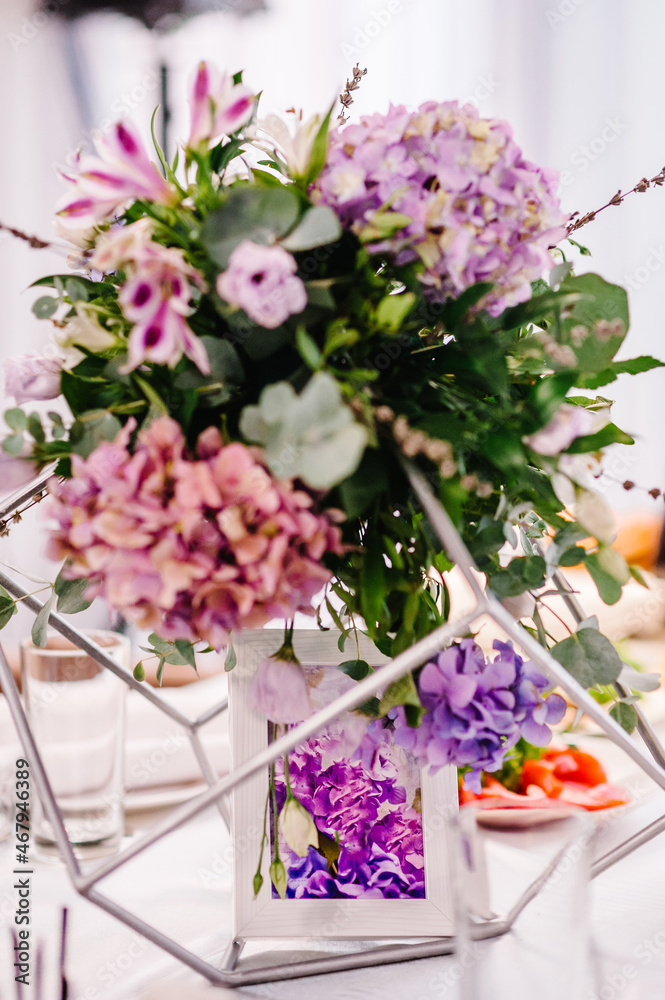 Festive table decorated with composition of violet, purple, pink flowers and greenery in the banquet hall. Table newlyweds in the area on wedding party.