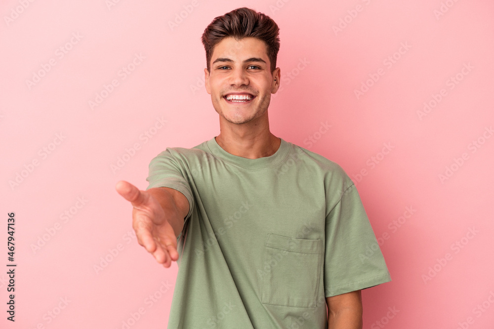 Young caucasian man isolated on pink background stretching hand at camera in greeting gesture.