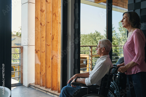 Elderly woman standing behind male wheelchair user near window