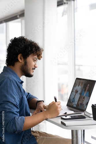 Focused young indian man listening interracial speakers watching lecture seminar webinar online and writing notes looking at laptop screen. Studying remotely or distant interview side view