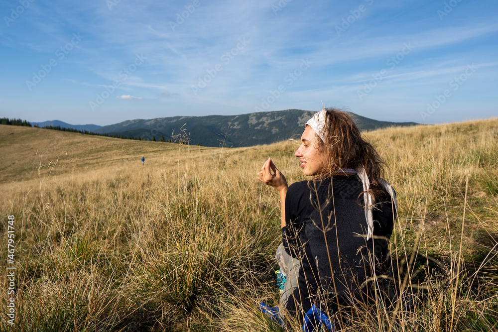 Rear view of a hiker woman dressed in a black hoodie standing on a grassy field in the mountains. Hot summer day in the mountains. Smiling female face. Rear view of a hiker woman dressed in a black hoodie standing on a grassy field in the mountains. Hot summer day in the mountains. Smiling female face.