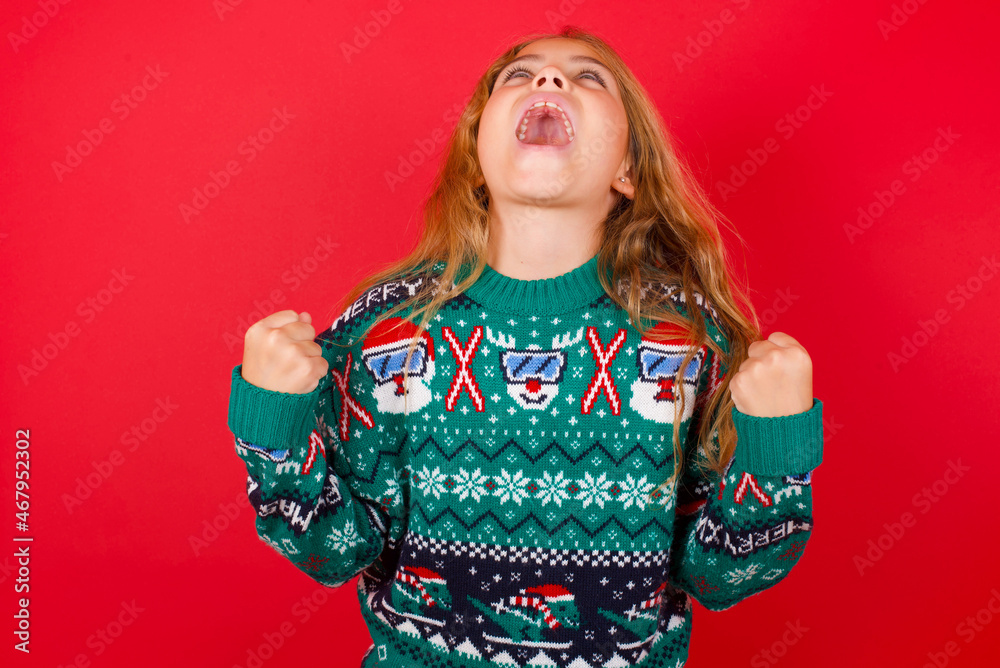brunette kid girl in knitted sweater Christmas over red background looks with excitement up, keeps hands raised, notices something unexpected. brunette kid girl in knitted sweater Christmas over red background looks with excitement up, keeps hands raised, notices something unexpected.