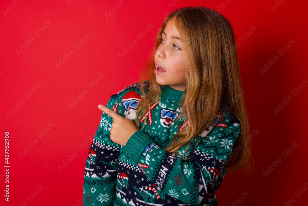 Happy cheerful smiling brunette kid girl in knitted sweater christmas over red background looking and pointing aside with hand. Copy space and advertisement concept. Happy cheerful smiling brunette kid girl in knitted sweater christmas over red background looking and pointing aside with hand. Copy space and advertisement concept.