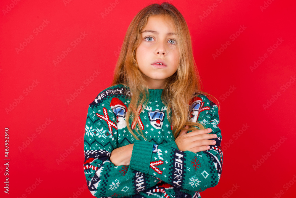 brunette kid girl in knitted sweater christmas over red background frowning his face in displeasure, keeping arms folded, waiting for an explanation. brunette kid girl in knitted sweater christmas over red background frowning his face in displeasure, keeping arms folded, waiting for an explanation.