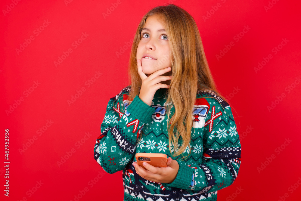 Image of a thinking dreaming brunette kid girl in knitted sweater christmas over red background using mobile phone and holding hand on face. Taking decisions and social media concept. Image of a thinking dreaming brunette kid girl in knitted sweater christmas over red background using mobile phone and holding hand on face. Taking decisions and social media concept.