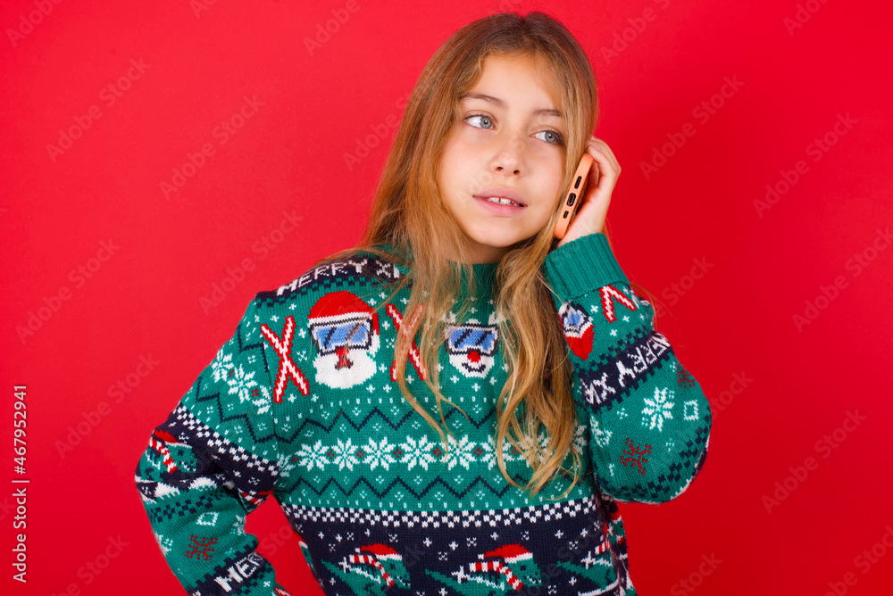 Portrait of successful joyful brunette kid girl in knitted sweater christmas over red background talking on mobile phone with friend. Lifestyle and communication concept Portrait of successful joyful brunette kid girl in knitted sweater christmas over red background talking on mobile phone with friend. Lifestyle and communication concept