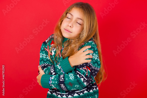 brunette kid girl in knitted sweater christmas over red background . Hugging oneself happy and positive, smiling confident. Self love and self care.