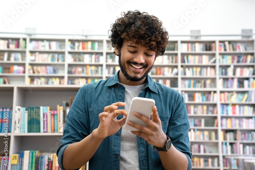 Overjoyed young Indian student chatting social media on smartphone in library