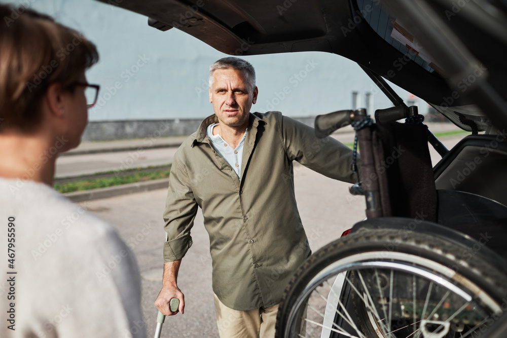 Portrait of mature couple unloading wheelchair out of car trunk in parking lot outdoors, copy space Portrait of mature couple unloading wheelchair out of car trunk in parking lot outdoors, copy space