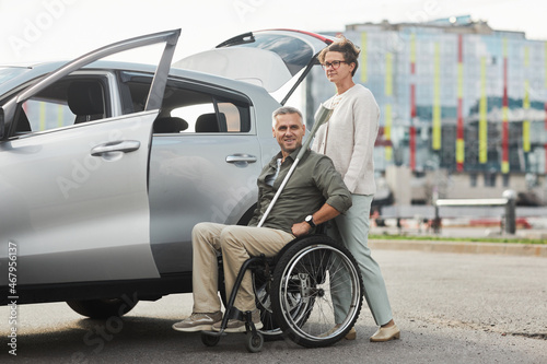 Full length portrait of mature couple with man in wheelchair by car in parking lot outdoors, copy space