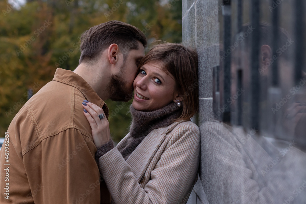 Fototapeta premium Young man kisses his beloved outside. She hugs him by the shoulders and smiles at the camera. Side view image