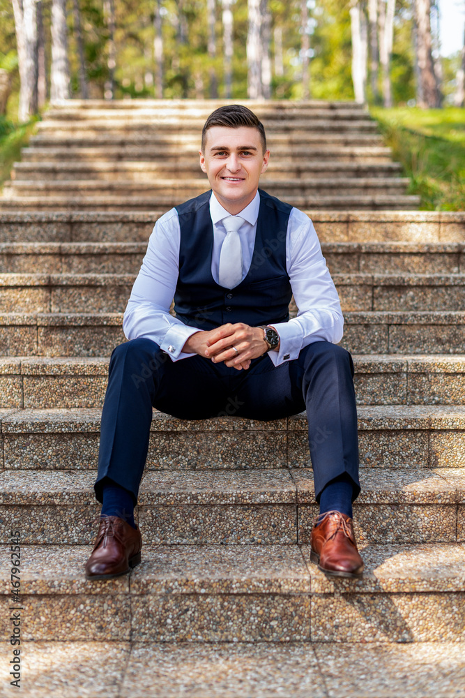 Handsome young groom wearing elegant and stylish dark blue suit outdoors portrait. Happy groom sitting on stairs looking and smiling at camera. Handsome young groom wearing elegant and stylish dark blue suit outdoors portrait. Happy groom sitting on stairs looking and smiling at camera.