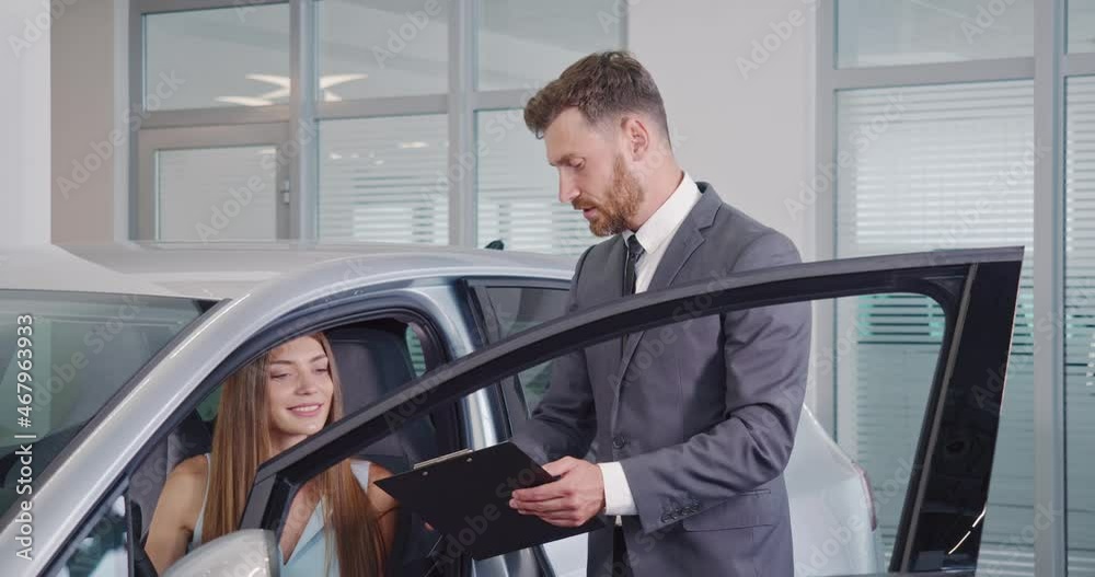 Professional car dealer in stylish suit with clipboard in hands showing female buyer new auto at salon. Happy caucasian woman sitting inside vehicle and examining leather interior. Professional car dealer in stylish suit with clipboard in hands showing female buyer new auto at salon. Happy caucasian woman sitting inside vehicle and examining leather interior.