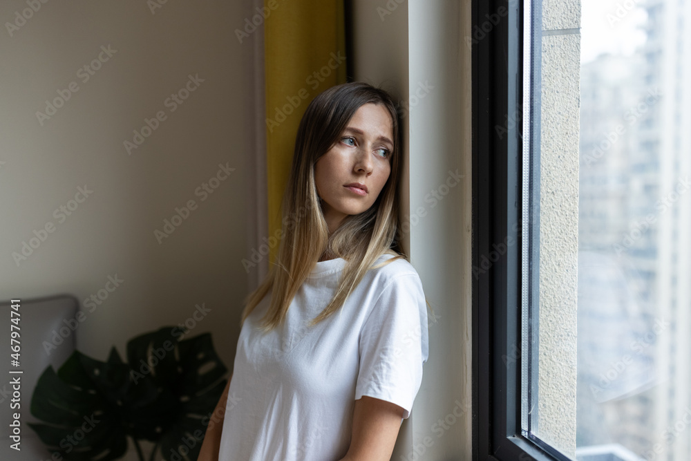 Unhappy young caucasian woman with blonde hair thinking about bad relationships problems, break up with boyfriend. Worried millennial girl standing in bedroom near window alone at home, suffering from