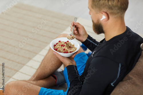Fototapeta Naklejka Na Ścianę i Meble -  Athlete enjoying healthy meal rich in fiber, protein and vitamins. Fit young man sitting on floor in living room, relaxing after fitness workout, eating natural vegetarian granola, listening to music