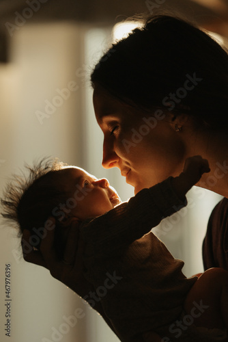 A close up shot of the happy mother who is holding the newborn baby in her hands and hugging her at home. The smiling infant and the mom in the sunlight in front of the window in the evening.
