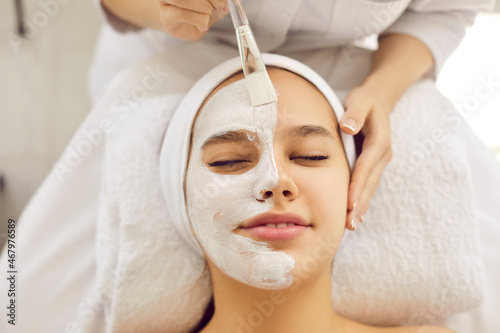 Cosmetologist applying calming cosmetic facial mask for fresh clear skin. Beautiful young woman with half face in kaolin clay lying eyes closed on soft towel in beauty salon or spa center, close up