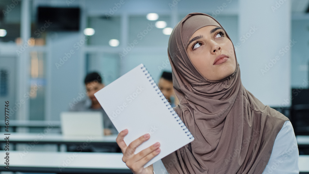 Pensive young Arabic female worker in hijab sitting at office desk using paper notebook making notes create new idea. Thoughtful millennial mixed race woman looking aside, pondering work planning Pensive young Arabic female worker in hijab sitting at office desk using paper notebook making notes create new idea. Thoughtful millennial mixed race woman looking aside, pondering work planning