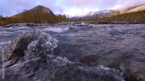 A clear stream running over stone boulders in the Khibiny. Abundant river in the mountains flows along the stone bottom in slow motion. Snow-capped mountain peaks in the background. 