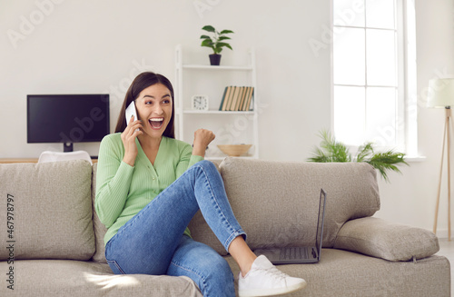 Young woman learns cool news while talking on phone. Beautiful Caucasian girl sitting on sofa with laptop at home gets super happy and excited when friend calls on cellphone and breaks wonderful news © Studio Romantic