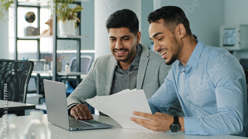 Bild auf Leinwand Cheerful positive friendly businessmen partners colleagues holding document contract looking at laptop screen discuss ideas having fun during break in office