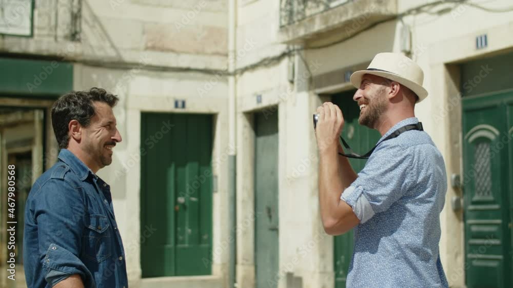 Side view of gay in hat taking photo of lover on camera outdoor. Medium shot of bearded Caucasian man posing for camera, smiling, taking picture during travel in Europe. LGBT, photoshoot concept Side view of gay in hat taking photo of lover on camera outdoor. Medium shot of bearded Caucasian man posing for camera, smiling, taking picture during travel in Europe. LGBT, photoshoot concept