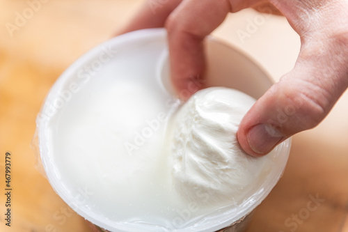 Macro closeup of hand holding fresh burrata mozzarella Italian cheese in storebought plastic container with brine and white color