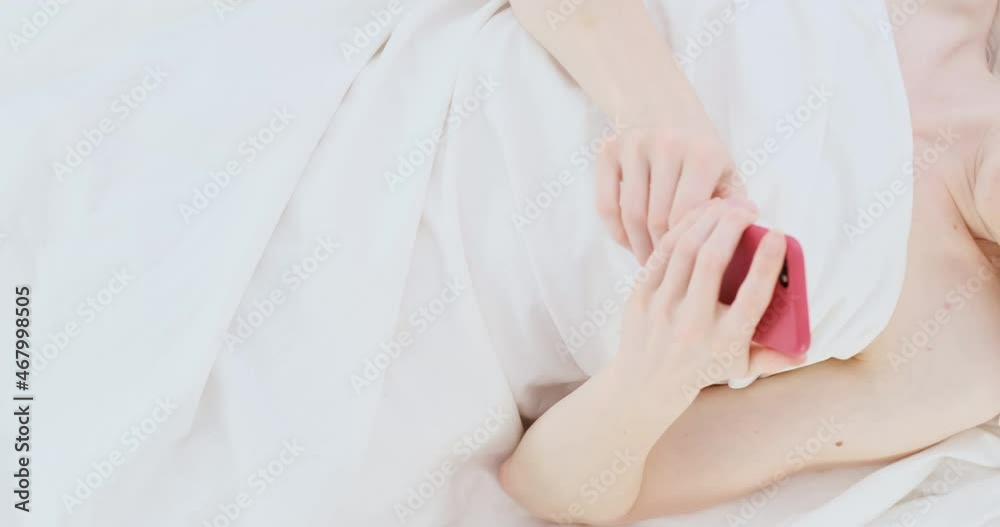 Young woman with long blond hair lies in bed and corresponds on the phone. Text messages in bed on white sheets. Young woman with long blond hair lies in bed and corresponds on the phone. Text messages in bed on white sheets.