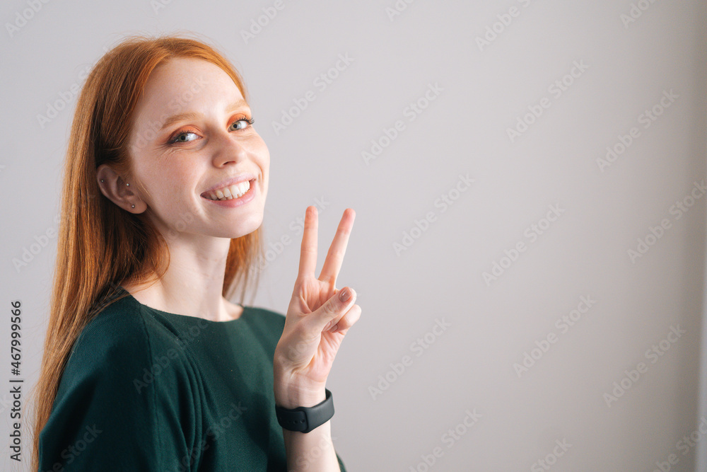Portrait of smiling redhead young woman model showing victory sign with fingers looking at camera on white background. Medium shot of red-haired lady with natural beauty posing in professional studio. Portrait of smiling redhead young woman model showing victory sign with fingers looking at camera on white background. Medium shot of red-haired lady with natural beauty posing in professional studio.
