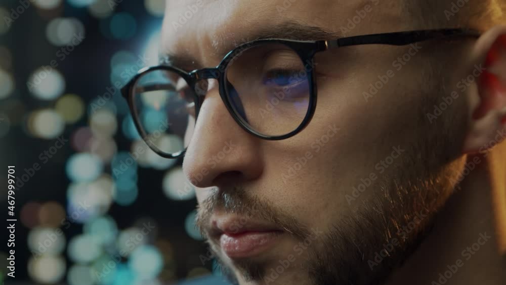 Close-up portrait of a young man working at the computer. Laptop display is reflected in glasses. Night city lights on the background. Business, coding and exchange concept. Close-up portrait of a young man working at the computer. Laptop display is reflected in glasses. Night city lights on the background. Business, coding and exchange concept.