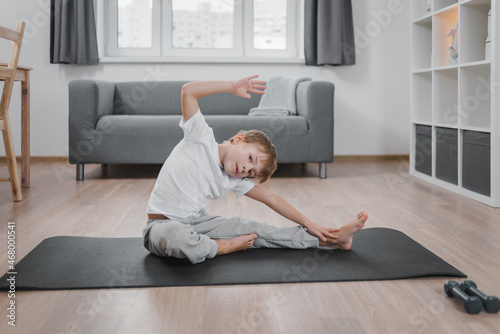 Caucasian child doing sports exercises at home on the living room floor. Dressed in home clothes: white T-shirt and gray trousers. The concept of promoting children's health through fitness.