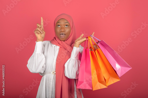 Portrait of a beautiful and excited African American girl wearing hijab looking and pointing up with finger and holding shopping bags isolated on a pink background
