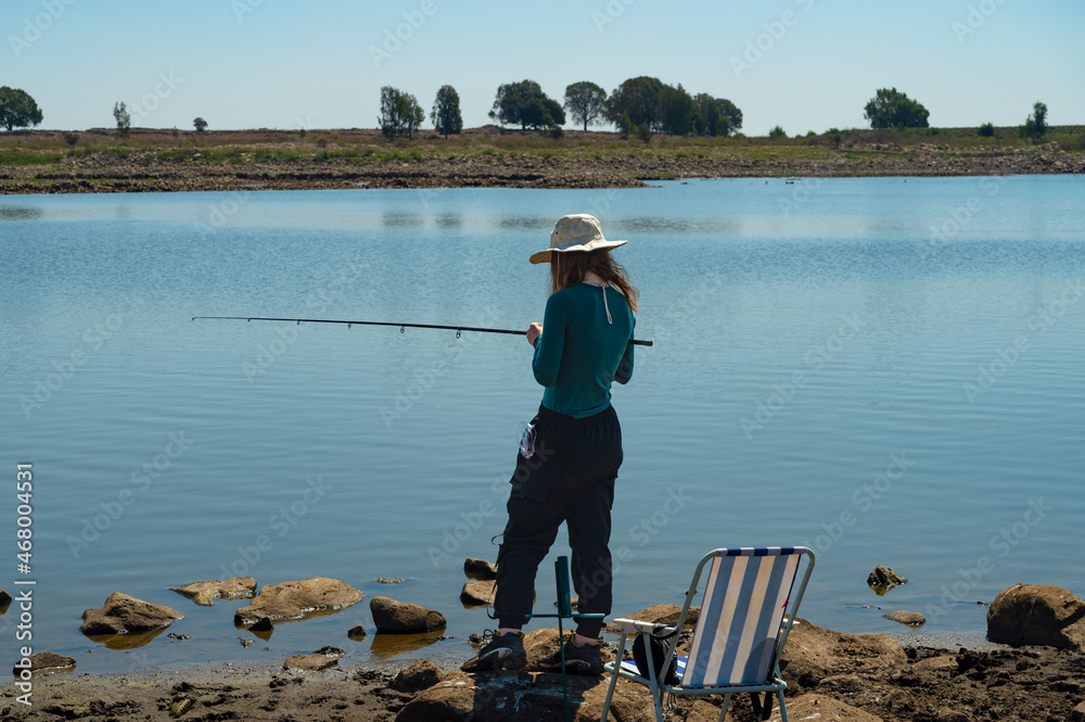 A girl in a straw hat with a fishing rod in her hand stands on the shore and poses