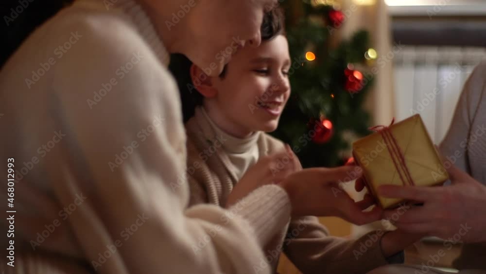 Close-up of happy child son giving Christmas present from mother and father sitting near xmas tree decorated balls and glowing garland. Festive family giving present box to loving kid at home. Close-up of happy child son giving Christmas present from mother and father sitting near xmas tree decorated balls and glowing garland. Festive family giving present box to loving kid at home.