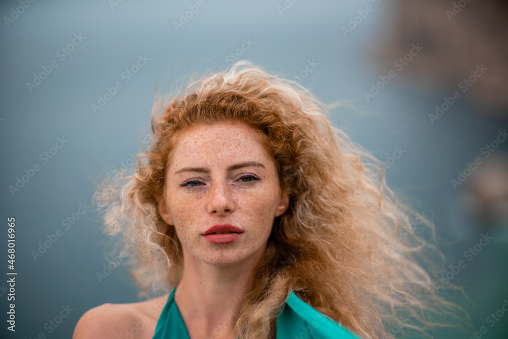 Outdoor portrait of a young beautiful natural redhead girl with freckles, long curly hair, in an emerald dress, posing against the background of the sea. Outdoor portrait of a young beautiful natural redhead girl with freckles, long curly hair, in an emerald dress, posing against the background of the sea.