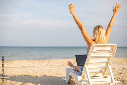 Beautiful young woman working on laptop when sitting in chaise-lounge on sandy beach