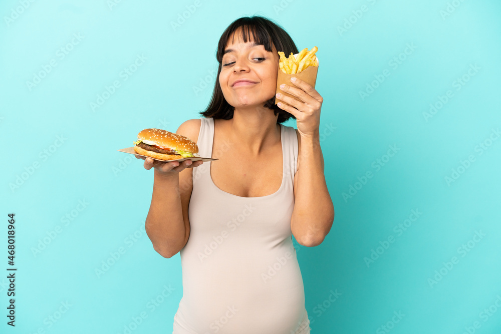 Young pregnant woman holding burger and fried chips over isolated blue background Young pregnant woman holding burger and fried chips over isolated blue background