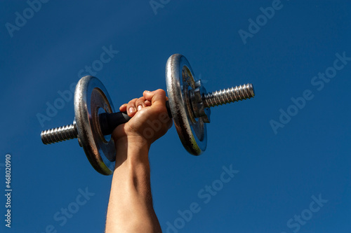 Adult lifting dumbbell with one hand with blue sky background. Outdoor fitness concept.