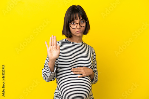 Young pregnant woman over isolated yellow background making stop gesture