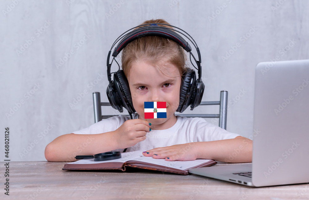 School female child with Dominican Republic flag. Child in headphones, with a book and laptop has lesson online School female child with Dominican Republic flag. Child in headphones, with a book and laptop has lesson online