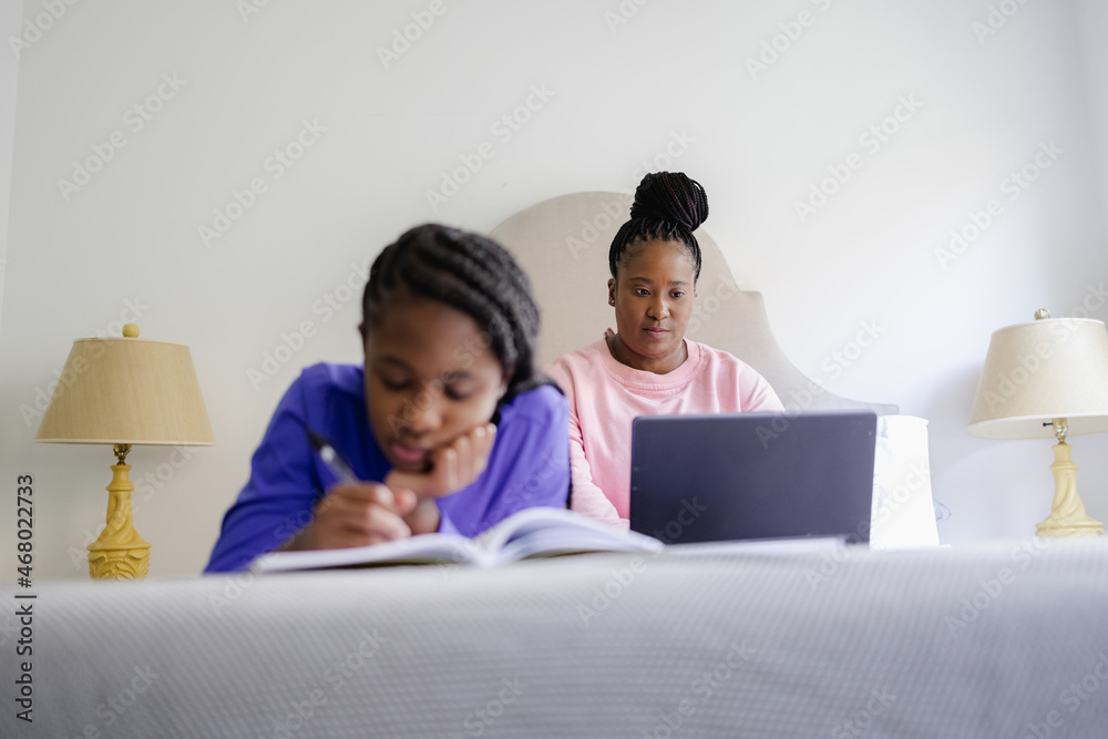 Teenage girl lying on bed and writing in notebook, mother using laptop in background Teenage girl lying on bed and writing in notebook, mother using laptop in background