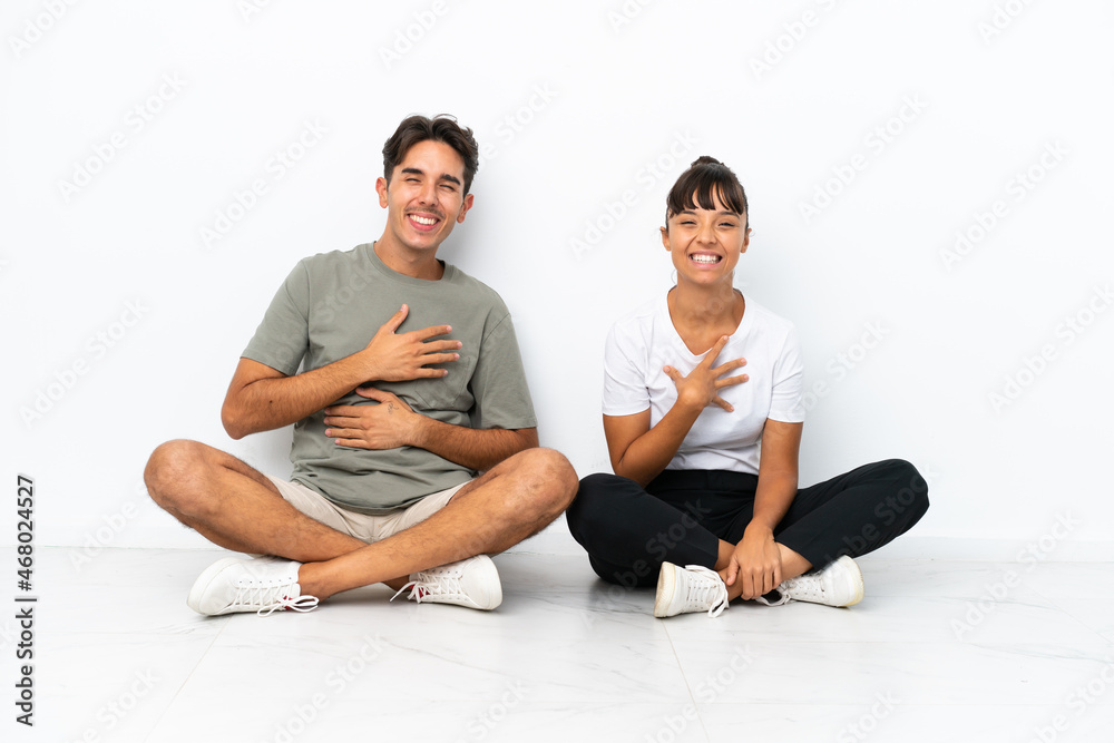Fototapeta premium Young mixed race couple sitting on the floor isolated on white background smiling a lot while putting hands on chest