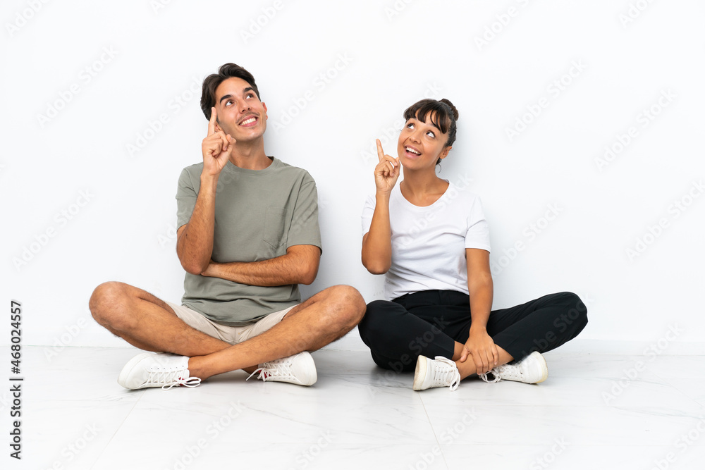 Young mixed race couple sitting on the floor isolated on white background pointing a great idea and looking up