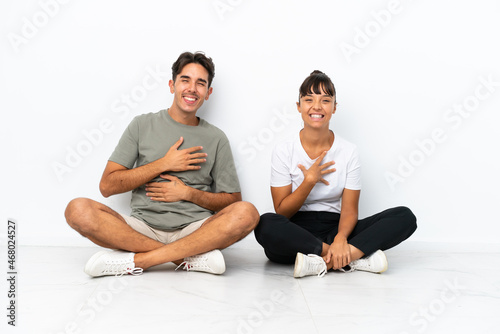 Young mixed race couple sitting on the floor isolated on white background smiling a lot while putting hands on chest